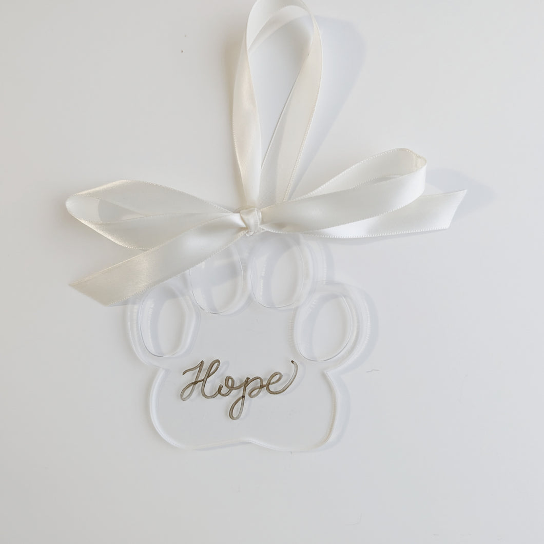 Clear paw shaped ornament with gold hand lettering, paired with a white ribbon to make a beautiful decoration on your holiday tree
