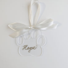 Load image into Gallery viewer, Clear paw shaped ornament with gold hand lettering, paired with a white ribbon to make a beautiful decoration on your holiday tree

