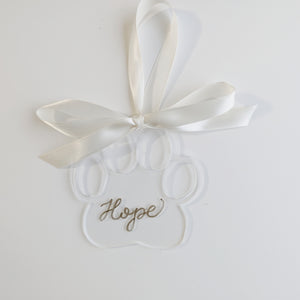 Clear paw shaped ornament with gold hand lettering, paired with a white ribbon to make a beautiful decoration on your holiday tree